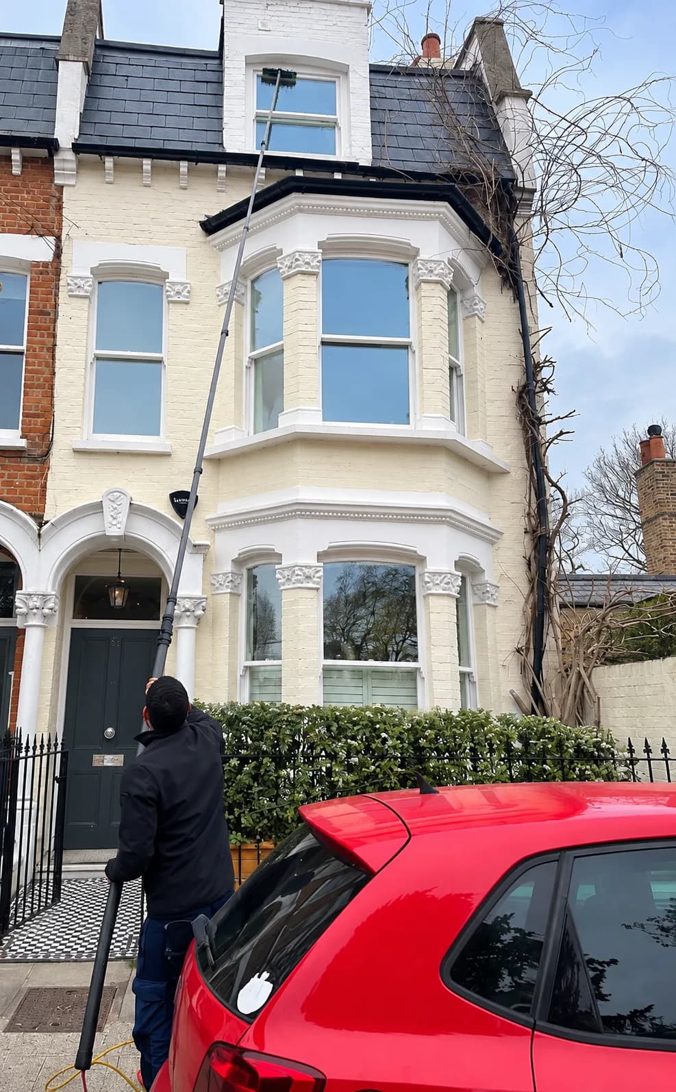 A technician cleaning the upper-floor bay windows of a cream Victorian terrace house with a water-fed pole.